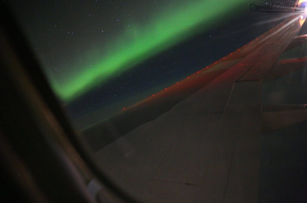 A green aurora arc seen from an aircraft window over the North Atlantic, the wing lit by red navigation lights