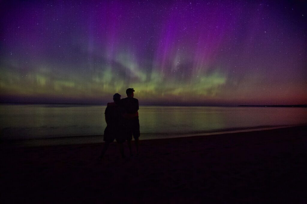 Two people on a Lake Michigan beach watching purple and green aurora rise over the water
