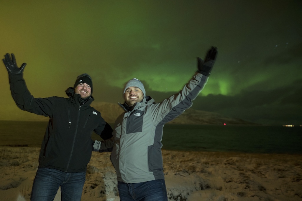 Two people with their arms raised beneath a bright green aurora on a beach in northern Norway