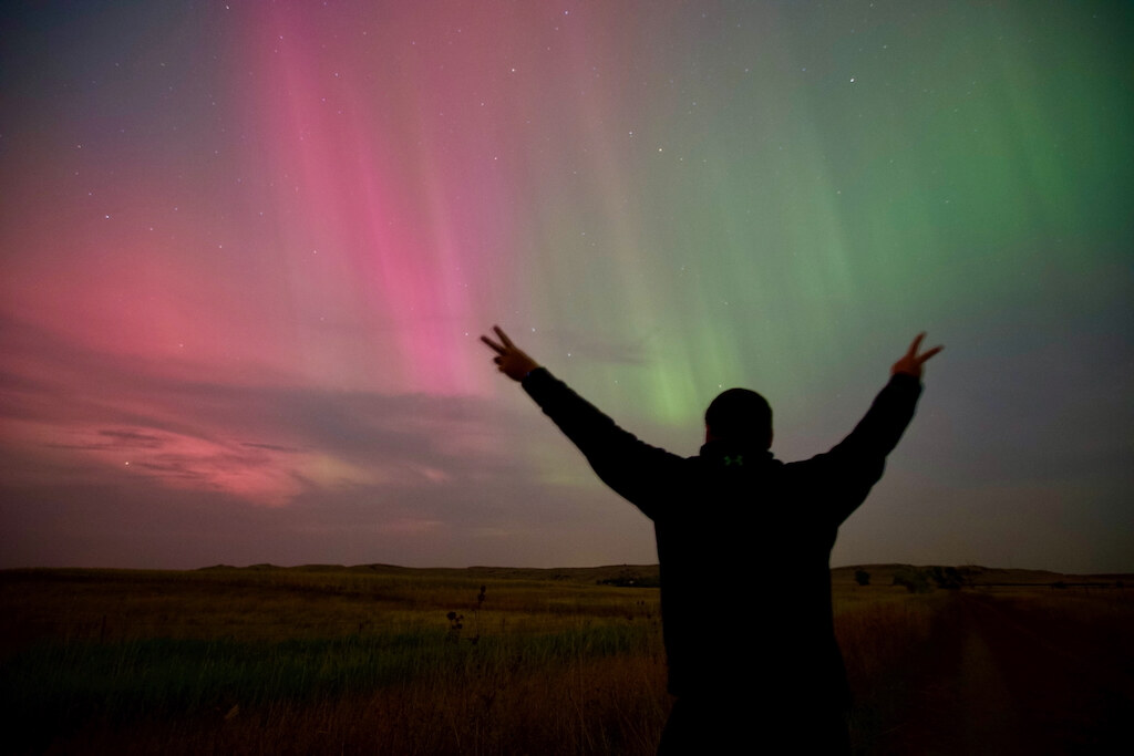 A person silhouetted with both arms raised beneath pink and green aurora over South Dakota prairie