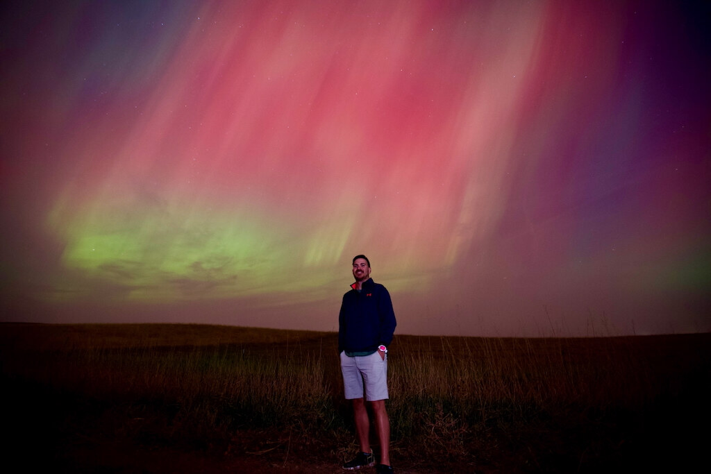 Daniel standing in prairie grass beneath a sky full of pink and green aurora in South Dakota