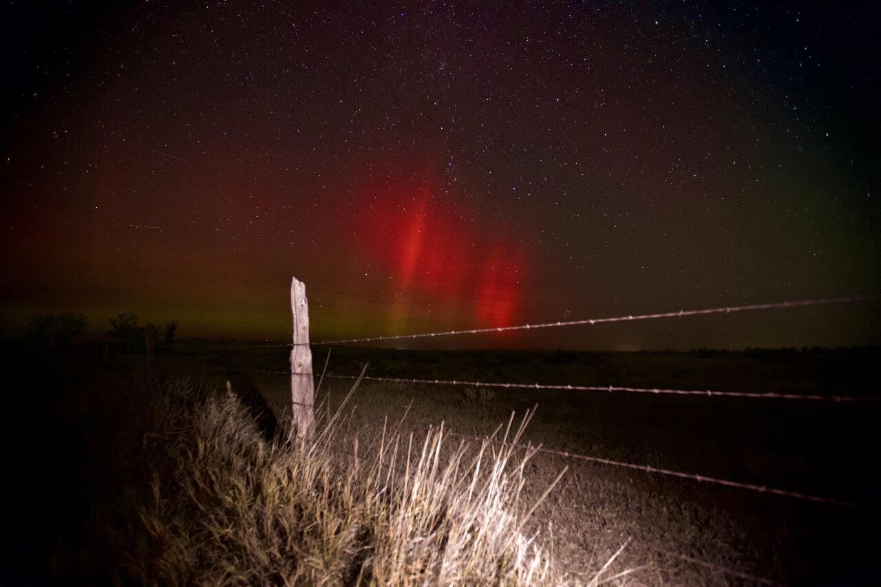 Red aurora pillars above a barbed wire fence and lit prairie grass in Wyoming