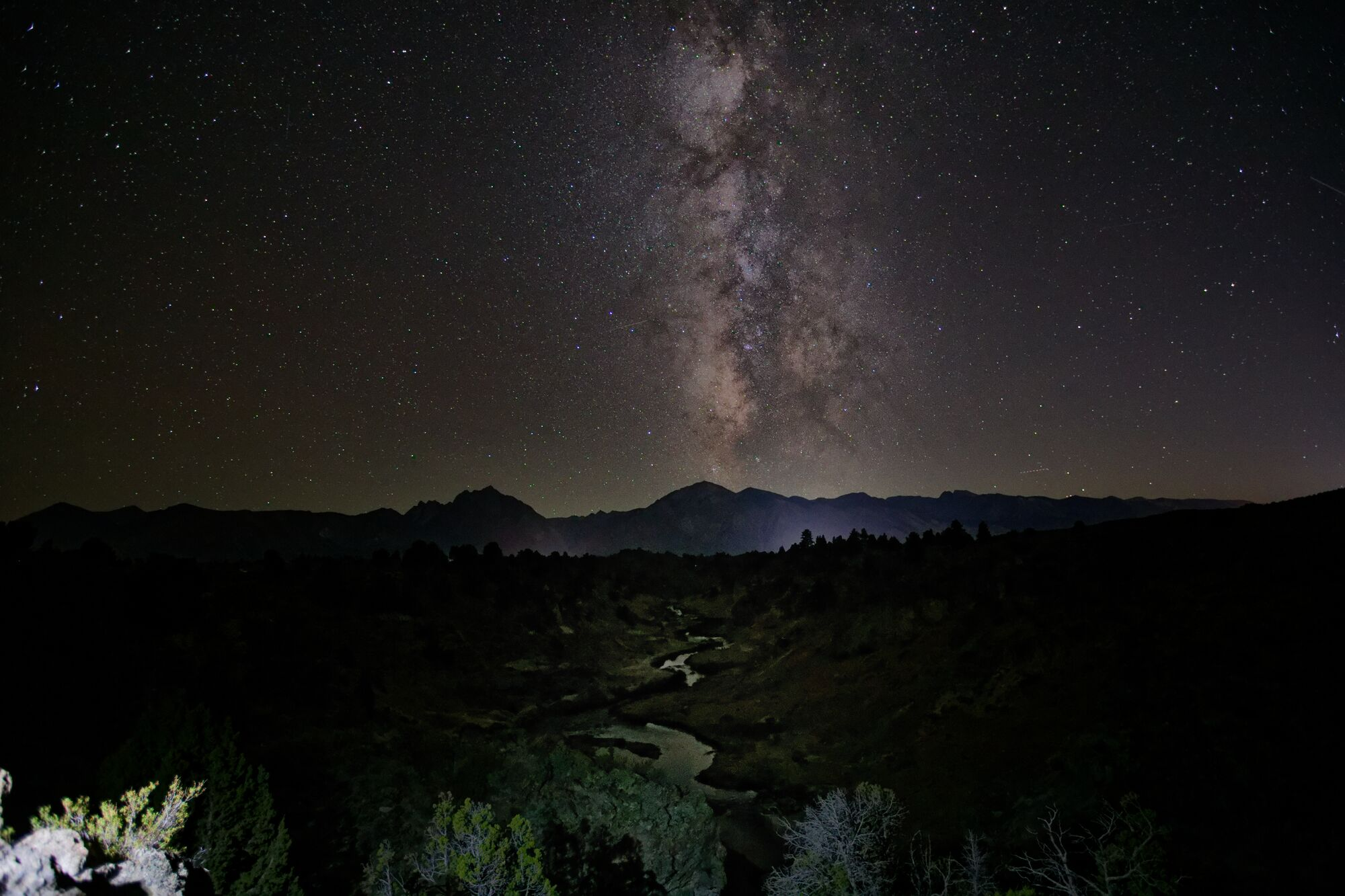 The Milky Way rising straight up above a dark mountain ridge, with a thin stream winding through the valley below at Hot Creek, California.