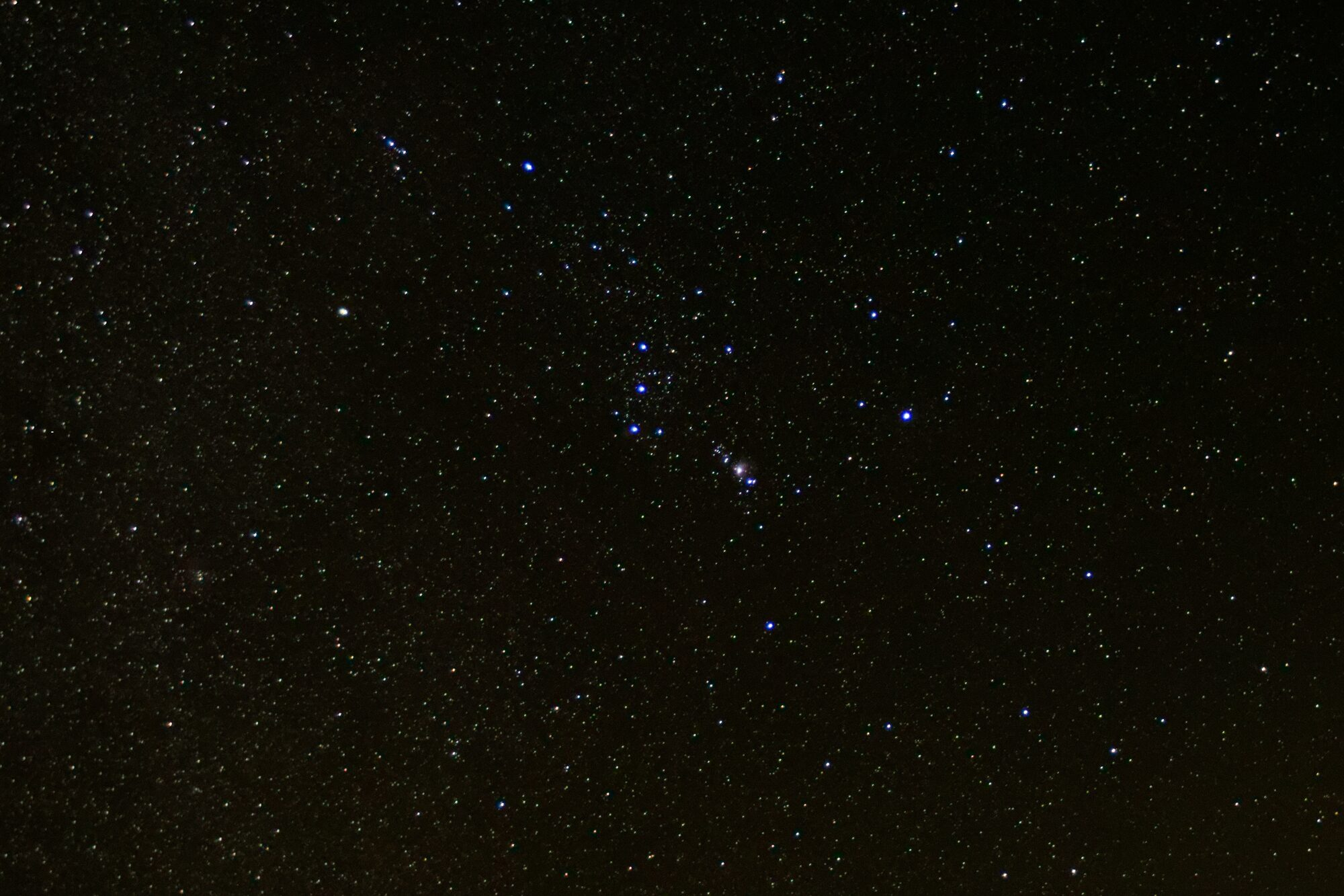 Orion tipped on its side in a dense field of stars, with the three Belt stars in a vertical line at center and the pink glow of the Orion Nebula just below and to the right.