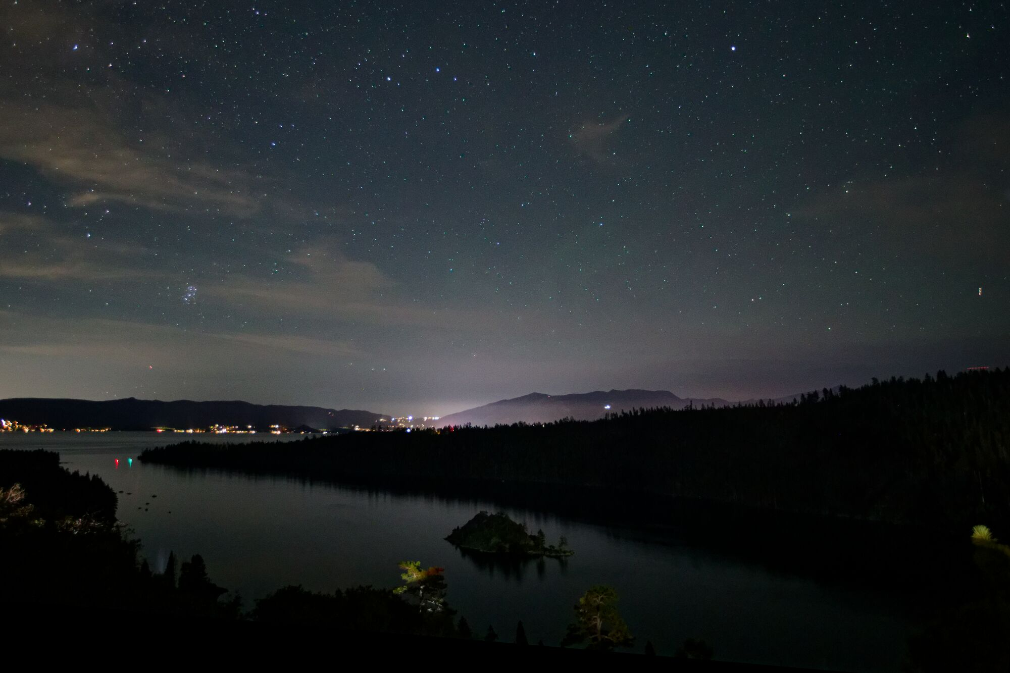 A tight cluster of blue-white stars above a dark ridge and the still water of Emerald Bay on Lake Tahoe, with thin clouds in the night sky.